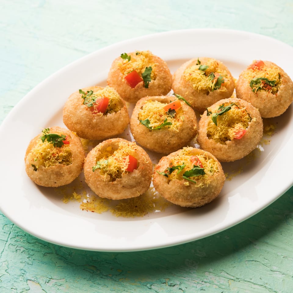 A white plate filled with nine golden pani puri, garnished with sev, cilantro, and diced tomatoes.