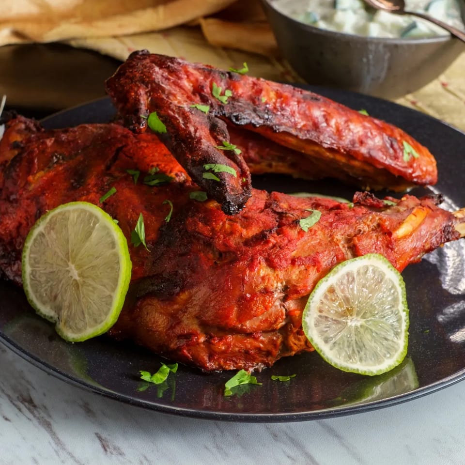 Two pieces of tandoori chicken, garnished with lime slices and cilantro, on a black plate.