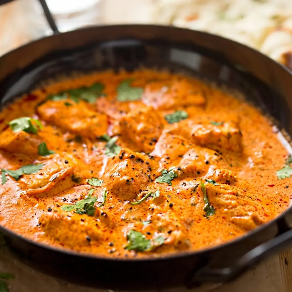 Close-up of butter chicken in a black pan, garnished with fresh cilantro and black pepper.